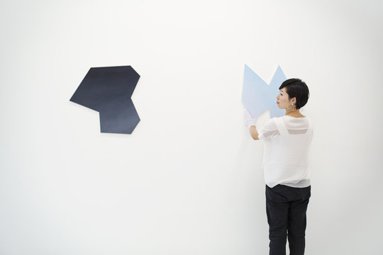Woman with  short black hair wearing white shirt hanging modern painting on white wall in art gallery.