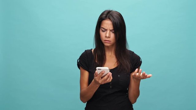 Confused brunette woman in t-shirt quarrels by smartphone over blue background