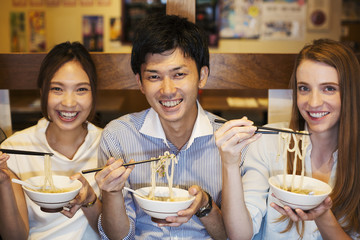Three smiling people sitting sidy by side at a table in a restaurant, eating from bowls using chopsticks.