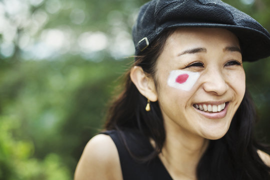 Portrait of smiling young woman with black hair wearing flat cap, Japanese flag painted on her cheek.