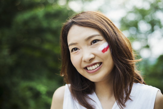 Portrait of young woman with brown hair, Japanese flag painted on her cheek, smiling at camera.