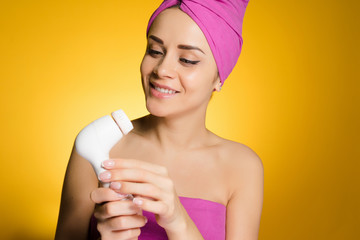smiling beautiful girl with a pink towel on her head holds an electric brush for deep cleaning of...