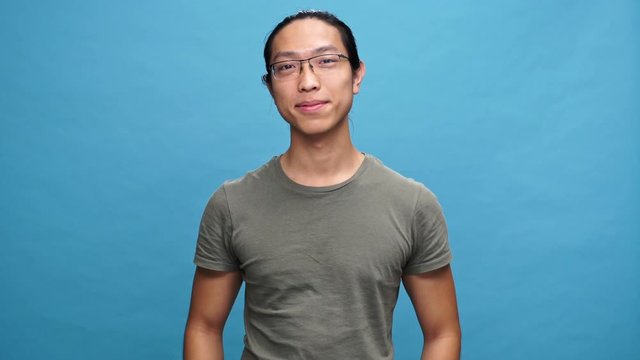Smiling asian man in t-shirt and eyeglasses showing thums up at the camera over blue background