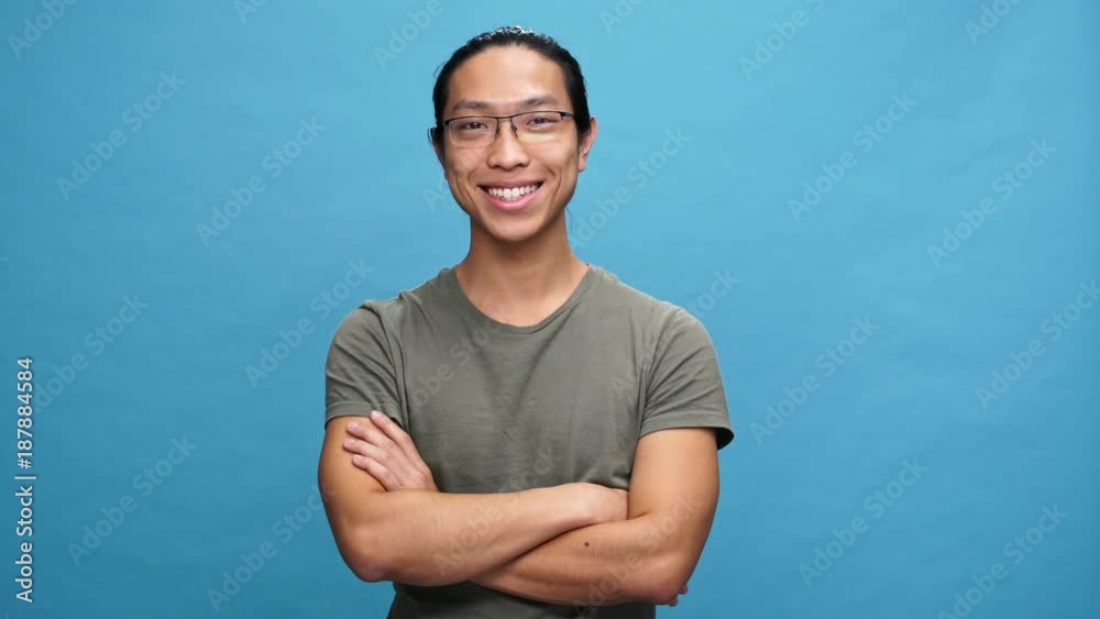 Sticker Happy asian man in t-shirt and eyeglasses holding crossed arms and agrees with something over blue background