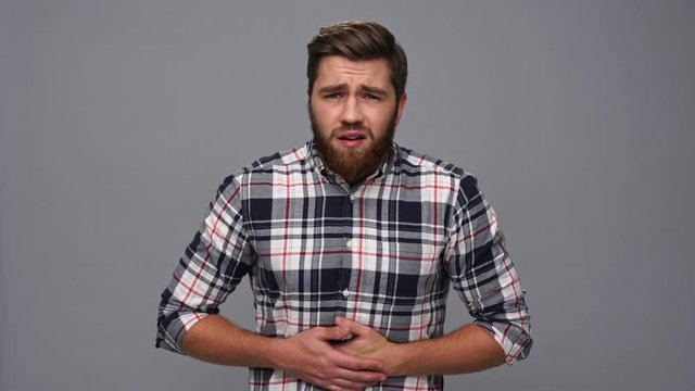 Confused bearded man in shirt having abdominal pain and touching his stomach over gray background
