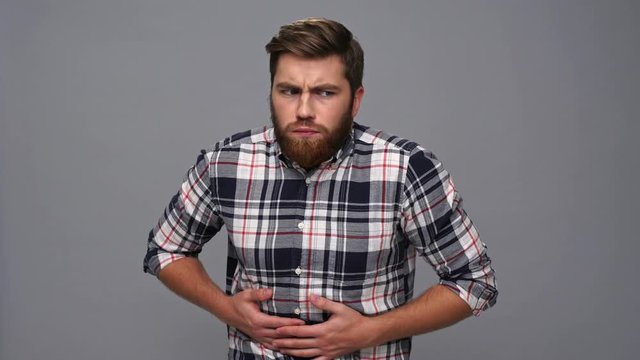 Confused bearded man in shirt having stomach upset and touching his stomach over gray background