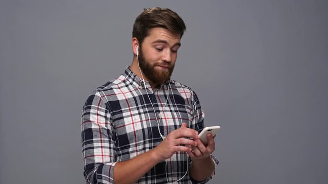 Smiling bearded man in shirt and headphones listening music on smartphone over gray background