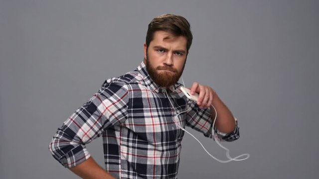 Funny bearded man in shirt and headphones listening music on smartphone and dancing over gray background