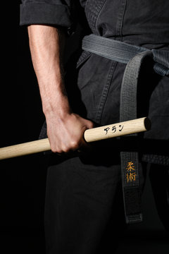 Black judo, aikido, or karate belt, tied in a knot with a hand holding a wood stick called Jô on black background