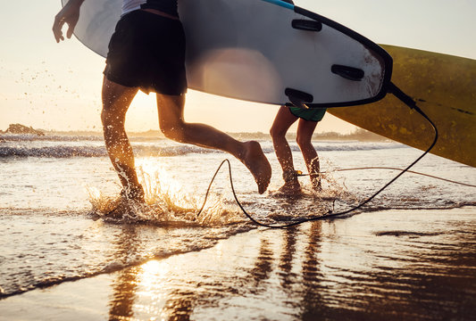 Son and father surfers run in ocean waves with long boards. Close up splashes and legs image