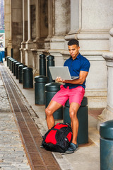 African American College Student travels, studies in New York. Man wearing blue short sleeve shirt, red shorts, with backpack on ground, sits on street, works on laptop computer. Filtered effect..