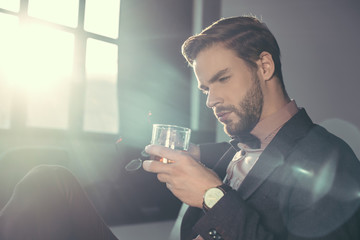 handsome bearded young man holding eyeglasses and glass of whisky
