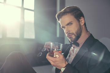stylish young man holding eyeglasses and glass of whisky, looking at camera