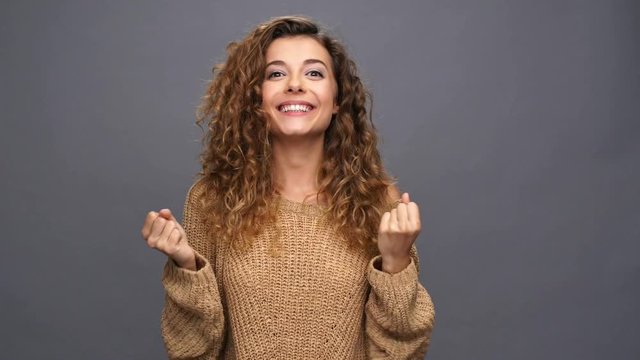 Surprised curly woman in sweater covering her mouth and rejoice over purple background