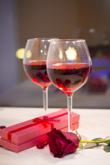 The concept of Love, Wedding, Proposal, Anniversary. A romantic evening with two glasses of red wine, a gift in a box and a beautiful red velvet rose. Dark blurred background
