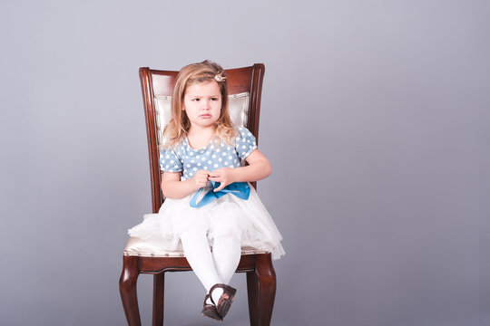 Sad kid girl 3-4 year old sitting on chair wearing dress and crown over gray background. Crying baby girl. Sadness.
