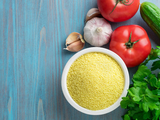 Vegetarian food - wheat, grain of raw couscous in a white bowl on a blue wooden background, fresh vegetables - parsley, cucumbers, tomatoes, top view