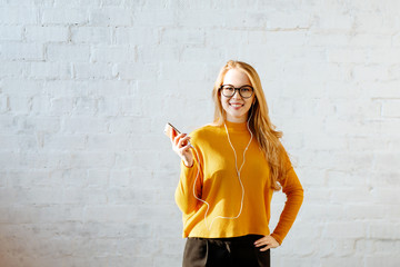Portrait of the young girl of the blonde with a yellow sweater against the background of a white brick wall. The girl smiles and listens to music on the smartphone in earphones. Copy space.