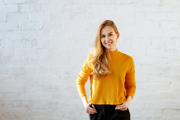 Portrait of the young girl of the blonde with a yellow sweater against the background of a white brick wall. The girl smiles and looks in the camera. Copy space.
