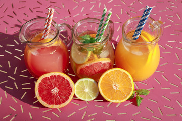 Three jugs with fresh drinks of grapefruit and orange juices, detox citrus water on an iron pink table.