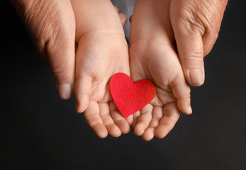 Hands of elderly man and baby with heart on dark background
