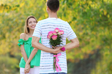 Young man hiding flowers and box with engagement ring before making proposal to his girlfriend in park