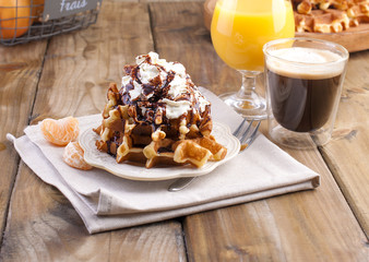 Sweet Belgian waffles for breakfast, decorated with white cream and chocolate sauce. A glass of coffee with black and orange juice, on a brown wooden background
