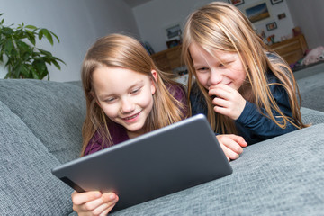 2 Mädchen liegen auf dem Sofa und schauen vergnügt auf den Bildschirm eines Tablets