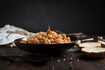 pilau in a brown ceramic dish, a kitchen towel, cherry tomatoes and a basket with spikelets of wheat, bread and a wooden spoon on a dark wooden table