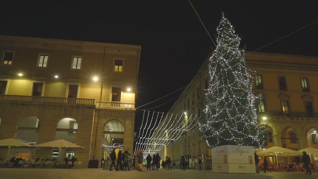 Tall Christmas tree in the middle of a square filled with people going by in the evening.