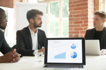 Project statistics on laptop screen with businessmen talk negotiate at background, executive team discussing project development work result, analyzing financial report charts graphs at meeting