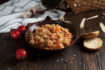 pilau in a brown ceramic dish, a kitchen towel, cherry tomatoes and a basket with spikelets of wheat, bread and a wooden spoon on a dark wooden table