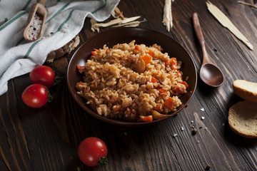 pilau in a brown ceramic dish, a kitchen towel, cherry tomatoes and a basket with spikelets of wheat, bread and a wooden spoon on a dark wooden table