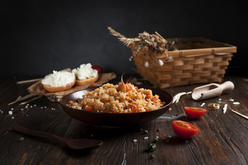 pilaf in a brown ceramic plate, cherry tomatoes and a basket with spikelets of wheat, sandwiches with cheese and a wooden spoon on a dark wooden table