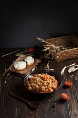 pilaf in a brown ceramic plate, cherry tomatoes and a basket with spikelets of wheat, sandwiches with cheese and a wooden spoon on a dark wooden table