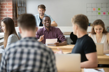 Smiling african-american businessman talking to colleague in coworking space, black attractive employee telling funny joke to coworker, friendly teamwork of co-working multiracial shared office
