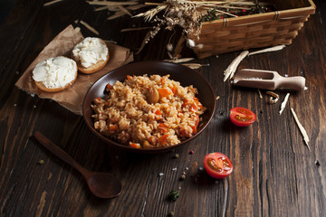 pilaf in a brown ceramic plate, cherry tomatoes and a basket with spikelets of wheat, sandwiches with cheese and a wooden spoon on a dark wooden table