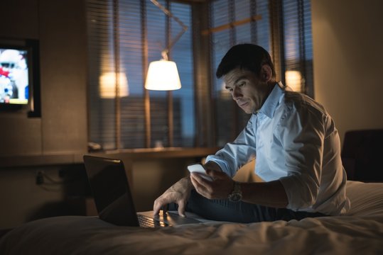 Businessman using laptop and mobile phone in bedroom