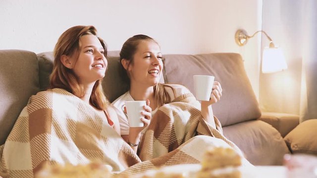 Two girlfriends are drinking tea in time watch TV at home.