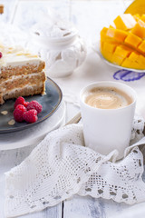 Cut the cake with white cream, for breakfast. A mango fruit. White background, tablecloth with lace, a cup of coffee and free space for text or advertising.