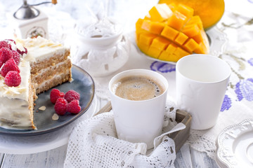 Cut the cake with white cream, for breakfast. A mango fruit. White background, tablecloth with lace, a cup of fragrant black coffee and free space for text or advertising.