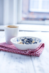 Plate with muesli and berries, coffee in a cup for breakfast. Napkin in a red cage on a white wooden table by the window. Free space for text.