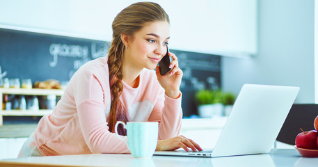 Young woman sits at the kitchen table using a laptop and talking on a cell phone