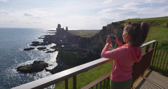 Tourist on travel taking photo with smart phone on Iceland of dramatic coast and ocean. Happy woman sightseeing taking pictures using smartphone visiting Arnarstapi, Snaefellsnes, West Iceland.