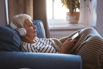 Senior woman listing to music on phone while siting on sofa in