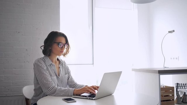 Side view of concentrated brunette woman in shirt and eyeglasses sitting by the table and using laptop computer indoors
