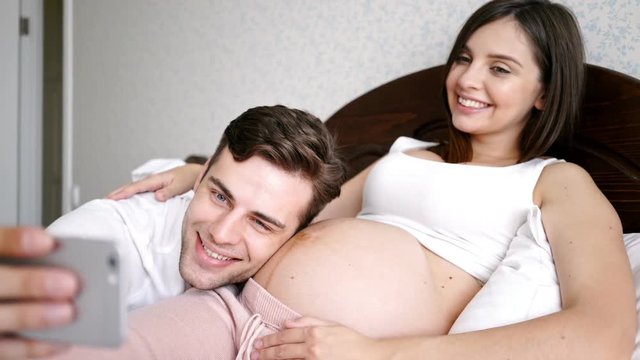 Funny young pregnant couple making selfie on smartphone while lying in bed