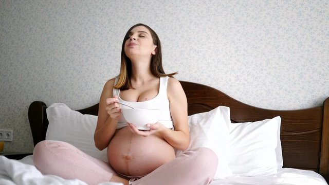Pleased pregnant woman eating food and enjoys it while sitting on bed at home
