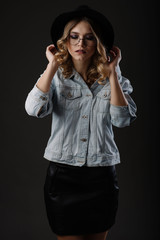 Fashion girl model posing on black background in the studio. Dressed in black hat, glasses and jeans jacket