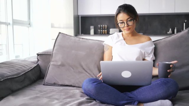 Smiling asian woman in eyeglasses sitting on sofa at home while using laptop computer and drinking coffee
